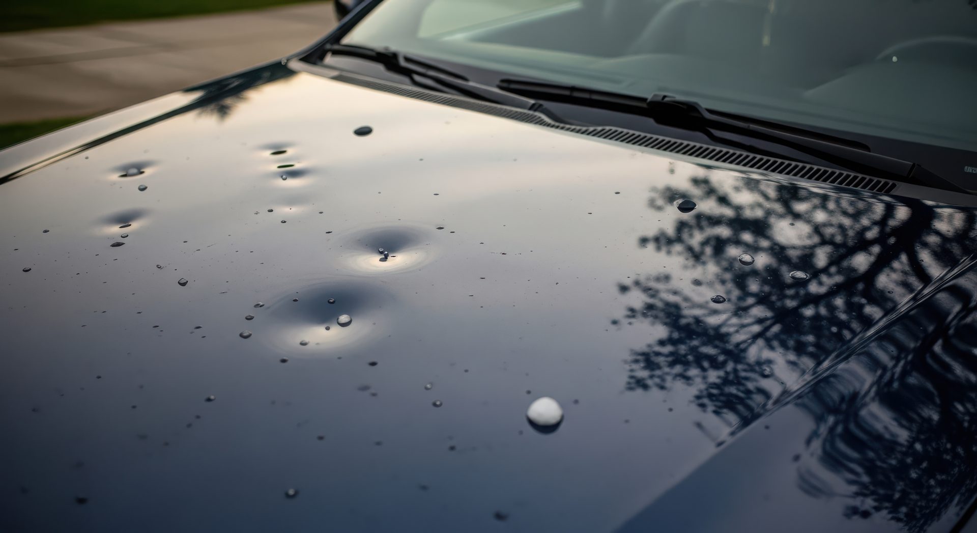 Hail dents across a black hood with a hailstone resting on the paint